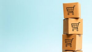 Four stacked cardboard boxes with shopping cart icons stand against a clean blue background, symbolizing e-commerce, delivery, packaging, and online retail.