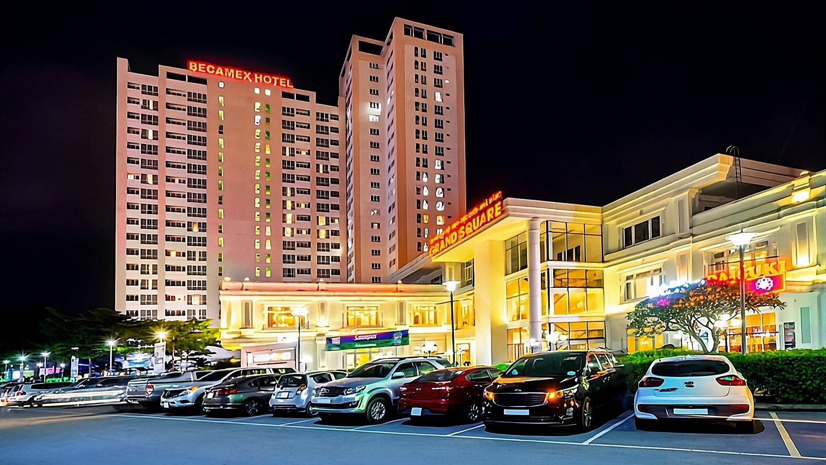 Nighttime exterior of Becamex Hotel with lit towers, Grand Square, and parked cars in the foreground.